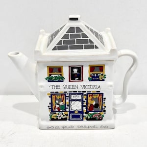 May include: White ceramic teapot shaped like a building, with a black and gray roof and the text "THE QUEEN VICTORIA" on the front. The teapot has a handle and spout, and the words "a pub teapot" are written at the bottom.