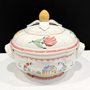 May include: A white ceramic soup tureen with a lid, featuring hand-painted floral and house motifs. The tureen has a pink trim and a yellow finial. It has two handles on the sides.