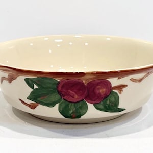 May include: Cream-colored ceramic bowl with a brown rim and a painted design of red fruit and green leaves. The bowl has a wide, shallow shape, suitable for serving.