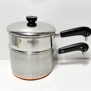 May include: A stainless steel double boiler with a copper-colored base. The unit features two stacked pots, a matching lid with a black knob, and two black heat-resistant handles, each equipped with a small metal hanging loop at the end.
