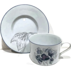 May include: A white porcelain teacup and saucer set with a blue trim. The saucer features a black and white line drawing of leaves. The teacup has a floral design with purple and green flowers.