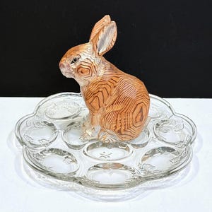 May include: A wooden rabbit figurine with detailed wood grain sits on a clear glass egg serving tray. The tray has a scalloped edge and individual egg compartments. The rabbit is light brown with darker brown graining.