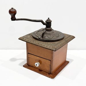 May include: An antique coffee grinder with a wooden body and a metal top and handle. The grinder has a small drawer with a white knob. The metal components have an ornate design. The handle is dark brown.