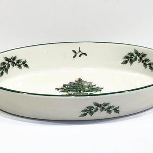 May include: White oval ceramic baking dish with a green rim and a green Christmas tree design. The dish is decorated with holly leaves and berries.