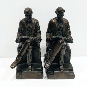 May include: A pair of bronze bookends featuring a seated figure of Abraham Lincoln reading a book. The bookends are detailed and have a realistic appearance.