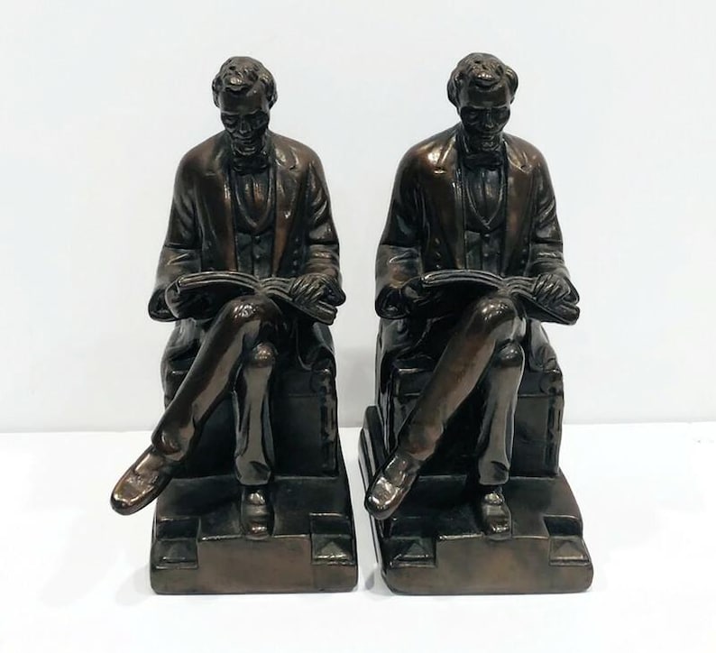 May include: A pair of bronze bookends featuring a seated figure of Abraham Lincoln reading a book. The bookends are detailed and have a realistic appearance.