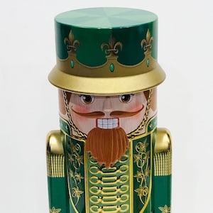 May include: A green and gold nutcracker tin with a whimsical design. The nutcracker is wearing a green hat with gold trim and a gold crown. The nutcracker's face is painted with a mustache and beard.