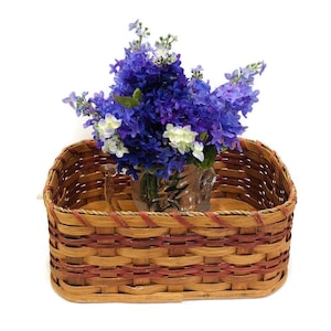 May include: A woven basket with a red and brown pattern, featuring a clear glass vase filled with a bouquet of purple and white flowers.