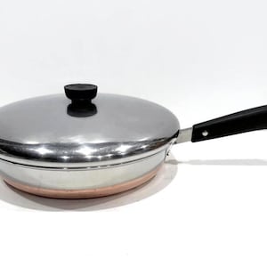 May include: A stainless steel frying pan with a lid. The pan has a copper-colored base and a black handle. The lid has a black knob. The pan is designed for cooking.
