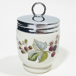 May include: White ceramic egg cup with a silver lid and a floral design featuring green leaves, white flowers, and red and black berries.