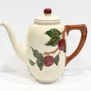 May include: Cream-colored ceramic teapot with a long spout and a brown handle. The teapot features a lid with a red knob and is decorated with a branch of red apples and green leaves.