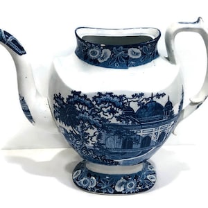 May include: A blue and white porcelain teapot with a floral design and a scene of a building with domes. The teapot has a wide base and a spout on the side.
