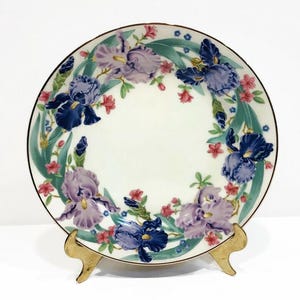 May include: A white ceramic plate with a gold rim features a floral design of purple, blue, and pink irises with green leaves. The plate is on a gold metal stand.