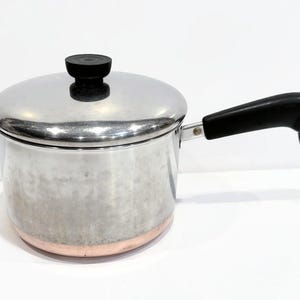 May include: A stainless steel saucepan with a copper-colored base and a matching lid. The pot features a black heat-resistant handle with a small metal hanging loop at the end and a black knob on the center of the lid for easy lifting and handling.