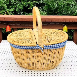 May include: A woven wicker picnic basket with a hinged lid and a sturdy handle. The basket is lined with blue and white gingham fabric. The basket is sitting on a white surface with black dots.