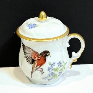 May include: White porcelain cup and lid with a gold rim and handle. The cup features a detailed painting of a bird with red and black feathers, and blue flowers. The lid has a gold knob and blue floral accents.