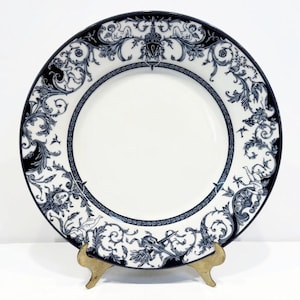 May include: White ceramic plate with a decorative dark blue and white floral and cherub design around the rim. The plate is displayed on a gold-colored stand. The plate has a wide rim with intricate patterns.