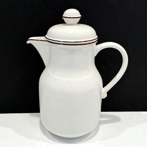 May include: A white ceramic coffee pot with a lid and handle. The pot has a rounded body, a spout, and a dark brown trim around the lid and spout. The lid has a small, round knob on top. The background is black.