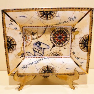 May include: A square ceramic dish with a gold trim and a compass design. The dish is on a gold metal stand. The dish features a compass rose and other nautical symbols.