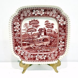 May include: A square, antique plate with a red and white floral and landscape design. The plate features a detailed landscape scene with a bridge and trees. The border has a floral pattern and a ruffled edge. The plate is displayed on a gold-colored stand.