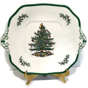 May include: A white square serving platter with a green border and a green Christmas tree design in the center. The tree is decorated with ornaments and presents. The platter has two handles on the sides.