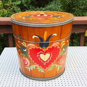 May include: A wooden storage container with a lid, painted with a traditional folk art design. The container is decorated with red, blue, green, and yellow flowers and hearts. The container is sitting on a white surface.