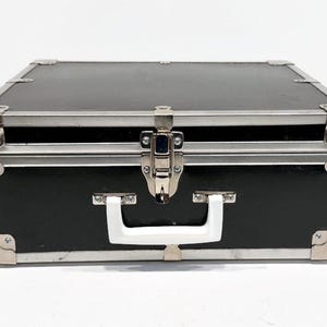 May include: Black rectangular case with silver metal trim and corner protectors. The case has a white handle and a silver latch. The case is closed and sits on a white surface.
