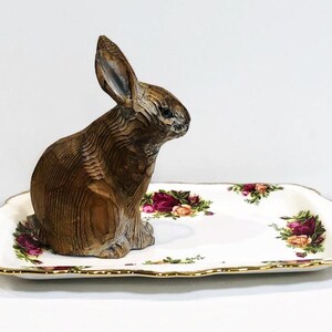 May include: A wooden rabbit figurine sits on a white porcelain serving tray with a floral pattern. The tray has a gold rim and features red, pink, and green roses.