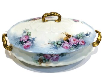 May include: A vintage porcelain serving dish with a lid. The dish is decorated with hand-painted pink roses and green leaves on a white background with a blue border. The lid has a gold handle and gold accents on the edges.