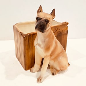 May include: A ceramic figurine of a Boxer dog with a tan coat and black muzzle, sitting next to a brown planter. The planter has a wood-grain texture and the word "PAL" is printed on the side. The dog is posed with its ears perked up.