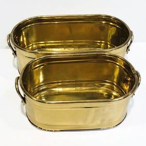 May include: Two polished brass oval basins with smooth, reflective surfaces and rounded edges. Each basin features two small, curved metal handles attached to the sides with rivets. The containers are nested, with one positioned behind the other.
