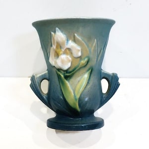May include: A matte blue ceramic vase featuring a raised relief of a white iris flower with green leaves. The vessel has a wide, flared rim, a circular base, and two symmetrical handles extending from the sides, finished in a muted blue tone.