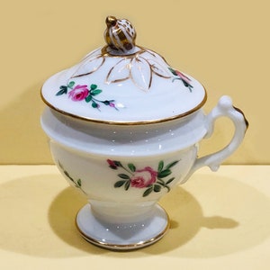 May include: White porcelain sugar bowl with lid, featuring pink rose floral designs and gold trim. The handle and lid finial are also accented with gold. A decorative piece for serving sugar or other condiments.