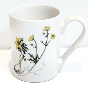 May include: A white ceramic mug featuring a botanical illustration of yellow buttercup flowers with green stems and leaves. The design shows the plant with roots visible at the base, rendered in a detailed, realistic style on the front surface.