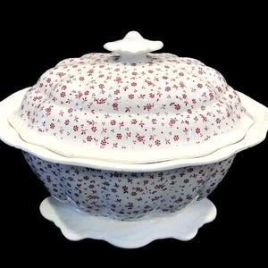 May include: A white ceramic tureen with a scalloped edge and a matching lid, featuring a repeating pattern of small dark red floral sprigs across the entire surface. The bowl sits on a pedestal base and includes a decorative knob on the lid.