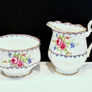 May include: A white porcelain creamer and sugar bowl set. The pieces feature a floral cross-stitch design in pink, red, blue, and green. Gold trim accents the rims and bases. The creamer has a curved handle and spout.