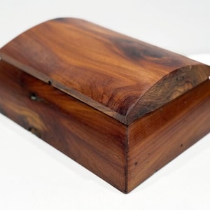 May include: A wooden box with a curved lid. The box is made of a dark wood with a natural finish. The lid is slightly raised and has a smooth surface.
