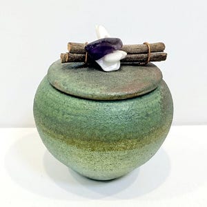 May include: A round, green ceramic jar with a matching lid. The lid is decorated with two brown wooden twigs, a polished purple amethyst stone, and white shell-like pieces, all secured together with thin copper wire across the top center.