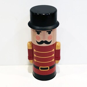 May include: A red and gold tin container shaped like a nutcracker. The nutcracker has a black top hat, a mustache, and a red and gold uniform.