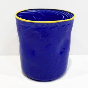 May include: A vibrant blue glass tumbler with a textured surface and a yellow rim. The handmade drinking glass has a unique, irregular shape, adding to its artistic appeal. The interior of the glass has a swirling pattern.