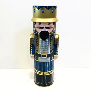 May include: A blue and gold nutcracker tin with a festive design. The nutcracker is wearing a blue uniform with gold trim and a gold crown. The tin is perfect for storing holiday treats or other small items.