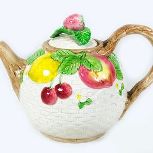 May include: A white ceramic teapot with a brown handle and a lid decorated with a lemon, peach, cherries, and strawberries. The teapot has a woven pattern and is decorated with green leaves.