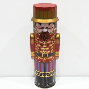 May include: A decorative, cylindrical nutcracker in the shape of a soldier. The nutcracker is primarily red with gold accents, a brown beard, and a red hat. The soldier's uniform features gold epaulets, a black belt, and black boots. The design is reminiscent of a traditional Christmas decoration.