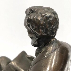 May include: Bronze sculpture of a man with a beard, wearing a suit jacket, sitting and reading a book. The sculpture is detailed and realistic.