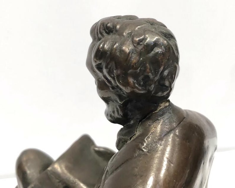 May include: Bronze sculpture of a man with a beard, wearing a suit jacket, sitting and reading a book. The sculpture is detailed and realistic.