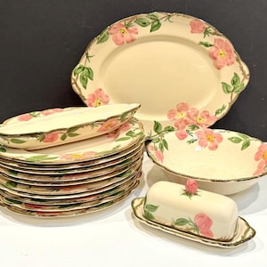 May include: A collection of cream-colored ceramic tableware featuring a floral design with pink flowers and green leaves. The set includes plates, a serving platter, a bowl, and a butter dish with a matching lid. The edges are trimmed with a gold-colored border.