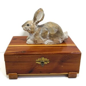 May include: A wooden box with a carved rabbit on top. The box is made of cedar wood and has a brass latch. The rabbit is painted white and has a natural wood grain.
