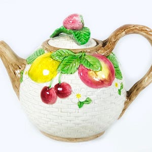 May include: A white ceramic teapot with a basket weave design, adorned with colorful fruit illustrations. The teapot features a brown handle and spout, and a strawberry-topped lid. The fruit includes cherries, a lemon, and a peach.