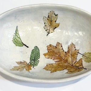 May include: Oval ceramic bowl with a white speckled finish. The bowl is decorated with detailed leaf designs in shades of green, brown, and gold. The leaves are arranged across the interior of the bowl.