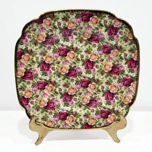 May include: A square ceramic serving dish with a gold rim and floral pattern. The dish is on a gold metal stand with three legs.
